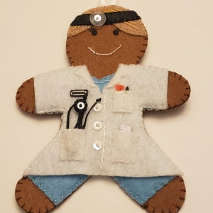 May include: A felt gingerbread person ornament dressed as a doctor, wearing a white coat, a blue shirt, a black headband, and a stethoscope. The ornament has a brown face with a smile and black eyes.