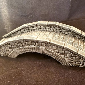 May include: A gray stone bridge model with a rounded arch and a textured surface. The bridge is made of small, irregular stones and has a rough, weathered look.