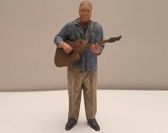 May include: A figurine of a person playing a guitar. The person is wearing a blue shirt and khaki pants. The guitar is brown and has a light brown pickguard.