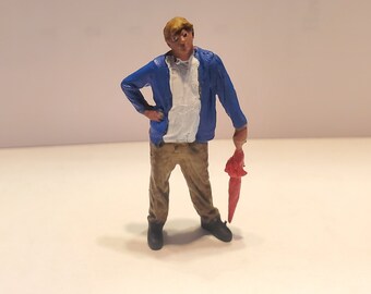 May include: A miniature figurine of a person wearing a blue jacket, white shirt, and khaki pants. The person is holding a red umbrella.
