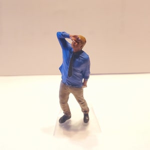 May include: A miniature figurine of a person wearing a blue shirt and khaki pants. The person is holding their hand to their forehead as if looking into the distance.