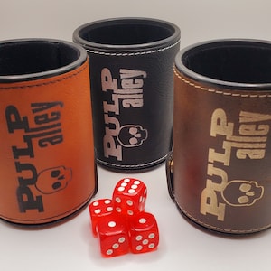 May include: Three leather dice cups with the text "Pulp Alley" and a skull graphic. The cups are brown, black, and orange. Four red dice are in front of the cups.