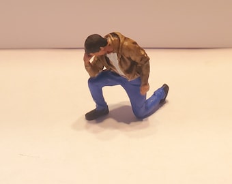 May include: A miniature figurine of a person kneeling on one knee, with their head bowed. The figure wears blue pants, a brown jacket, a white shirt, and black shoes. The figurine is set against a plain white background.