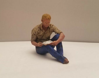 May include: A small plastic figurine of a person sitting with their legs crossed, wearing a brown shirt and blue jeans. The person is holding a piece of paper in their hands.