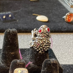 Puede incluir: Un anillo de plata con forma de rana con ojos de piedras preciosas rojas. El anillo se muestra en un soporte de anillo negro.