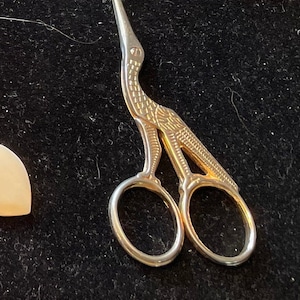 May include: Gold-toned scissors shaped like a bird with a pointed beak. The scissors are closed and have a decorative design.