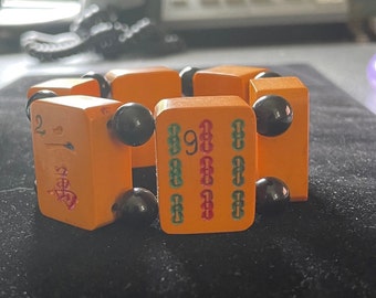 Puede incluir: Pulsera naranja y negra hecha de fichas de mahjong con cuentas negras. Las fichas tienen números y caracteres chinos. La pulsera está sobre una superficie negra.