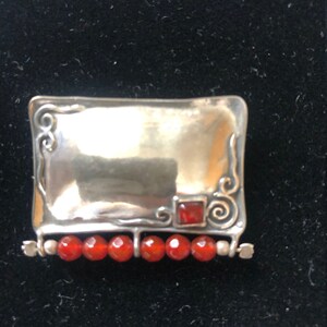 May include: A silver brooch with a rectangular frame and a red carnelian stone in the center. The frame is decorated with swirls and the brooch has a row of red carnelian beads.