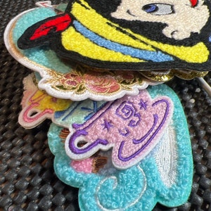 May include: A stack of colorful embroidered patches featuring a cartoon character, a floral design, and a teacup. The patches are made of soft, fuzzy fabric and have a variety of colors, including blue, pink, yellow, and green.