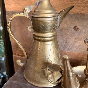 May include: A brass coffee pot with a tapered body, a pointed lid, and a curved handle. The surface features etched designs including geometric shapes and plant motifs. Beside it sits a small brass conical object with a circular ring attachment.