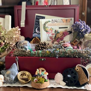 Puede incluir: Una caja de madera color burdeos llena de flores secas, una piña, una pequeña figura de hada y un quemador de incienso decorativo. Se ve una tarjeta con el texto "There is magic in the stars".