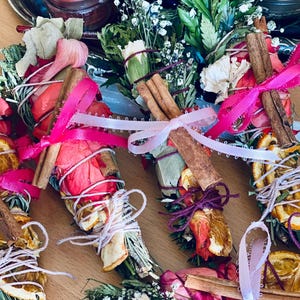 May include: A collection of five smudge sticks wrapped in pink and lavender ribbons. Each smudge stick is made with dried herbs, flowers, and spices, including cinnamon sticks, dried orange slices, and pink rose petals.