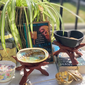 May include: A green spider plant in a woven basket sits on a table with a book titled "Like Water for Chocolate: A Novel in Monthly Installments with Recipes, Romances and Home Remedies" and a variety of decorative items, including a seashell, a small incense burner, and a gold incense holder.