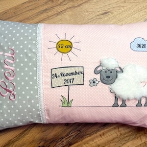 Op de afbeelding: Een roze en grijs babykussen met een witte kantversiering. Het kussen heeft een schaap, een zon, een wolk en een bord met de tekst "24. November 2017". Het kussen heeft ook de naam "Leni" en de afmetingen "52 cm" en "3620 g" erop geborduurd.