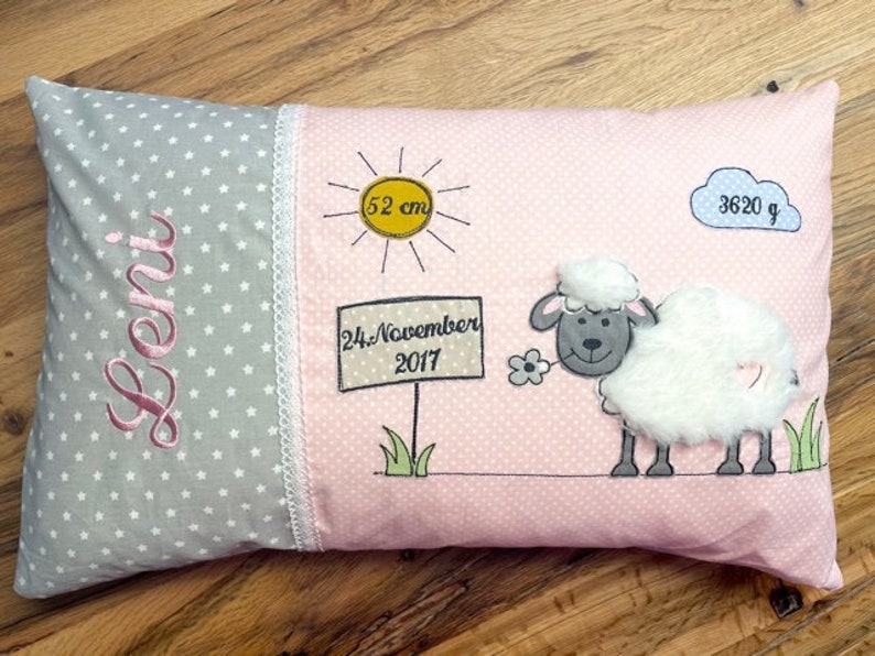 Op de afbeelding: Een roze en grijs babykussen met een witte kantversiering. Het kussen heeft een schaap, een zon, een wolk en een bord met de tekst "24. November 2017". Het kussen heeft ook de naam "Leni" en de afmetingen "52 cm" en "3620 g" erop geborduurd.