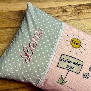 Op de afbeelding: Een roze en grijs babykussen met een witte kantversiering. Het kussen heeft een grijze achtergrond met witte sterren en de naam "Leni" geborduurd in roze. De andere kant van het kussen is roze met witte stippen en heeft een zon met de tekst "52 cm", een bord met de tekst "24. November 2017" en een cartoon schaap.