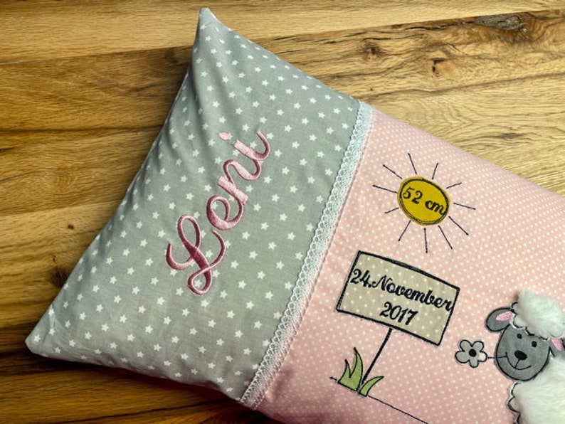 Op de afbeelding: Een roze en grijs babykussen met een witte kantversiering. Het kussen heeft een grijze achtergrond met witte sterren en de naam "Leni" geborduurd in roze. De andere kant van het kussen is roze met witte stippen en heeft een zon met de tekst "52 cm", een bord met de tekst "24. November 2017" en een cartoon schaap.