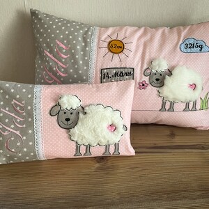 Op de afbeelding: Twee decoratieve kussens met een roze en grijze stof. De kussens hebben een wit schaap met een roze hart aan de zijkant en de naam "Jola" geborduurd in roze. Het grotere kussen heeft ook een zon, een wolk en de tekst "3215g" en "14. M&auml;rz" geborduurd erop.