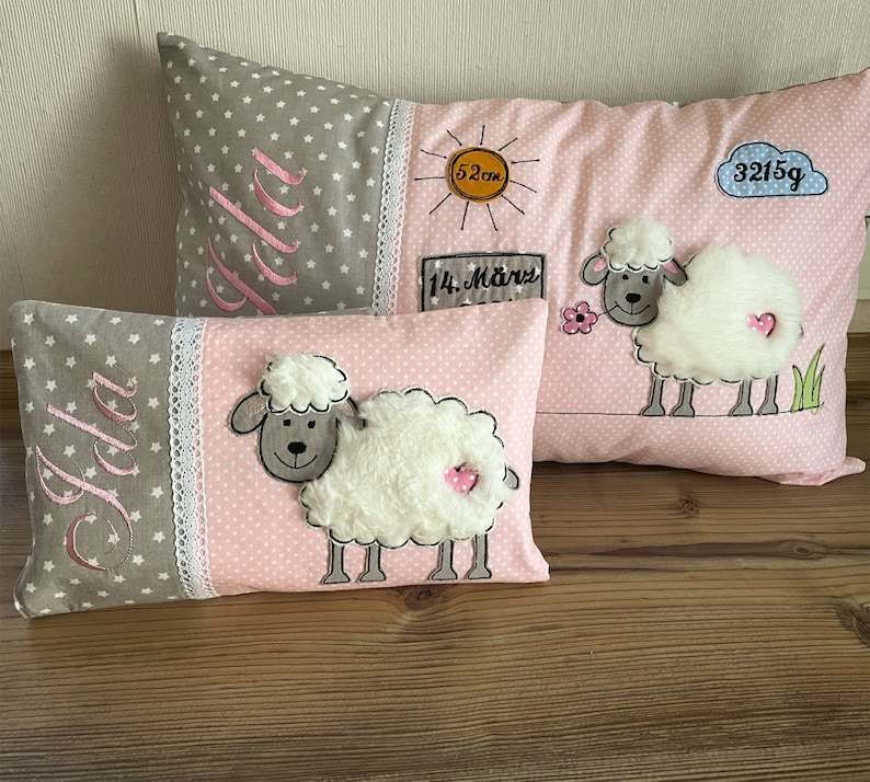 Op de afbeelding: Twee decoratieve kussens met een roze en grijze stof. De kussens hebben een wit schaap met een roze hart aan de zijkant en de naam "Jola" geborduurd in roze. Het grotere kussen heeft ook een zon, een wolk en de tekst "3215g" en "14. M&auml;rz" geborduurd erop.