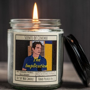 Op de afbeelding: Een brandende sojawas kaars in een glazen pot met een zwart deksel. Het etiket luidt "Scents of Cinema" en toont een illustratie van een man met de tekst "The Implication". De kaars is met de hand gegoten en heeft een geur van sandelhout, patchouli en bergamot.