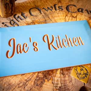May include: A light blue rectangular sign with the text "Jac's Kitchen" in gold lettering. The sign is on a wooden surface with the text "Night Owls Cave" carved into the wood.