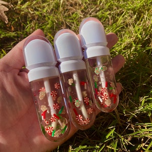 May include: Three clear tubes filled with a glittery, translucent liquid. The tubes have white caps and contain small, colorful, holiday-themed embellishments, such as candy canes, gingerbread men, and Christmas trees.