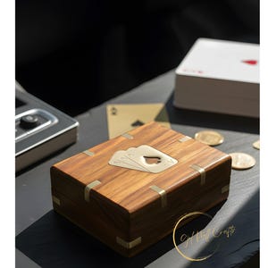 Könnte beinhalten: Holzspielkartenbox mit Messingakzenten und einem dekorativen Kartenspiel-Emblem. Ein Kartenspiel und Goldmünzen sind in der Nähe. Die Box hat eine satte braune Farbe und scheint aus Holz zu sein.