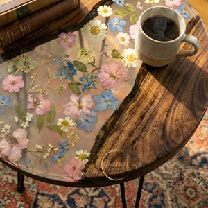 Może przedstawiać: Okrągły drewniany stolik kawowy z naturalną krawędzią. Stół ma przezroczysty żywiczny blat z kolorowymi suszonymi kwiatami. Na stole stoi kubek kawy i złoty pierścionek. Ma czarne metalowe nogi.