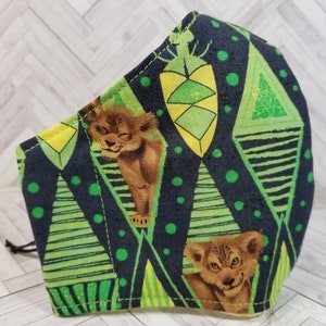 May include: A black face mask with a green and yellow geometric pattern featuring a lion cub design. The pattern includes green and yellow triangles, stripes, and dots.