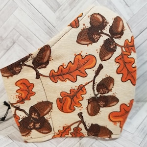 May include: A beige fabric face mask with a pattern of brown acorns and orange oak leaves.