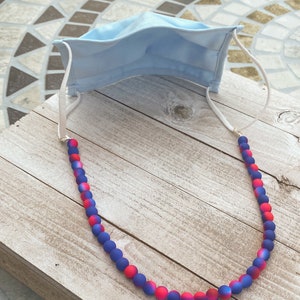 May include: A blue face mask with a white beaded chain. The beads are ombre red and blue.