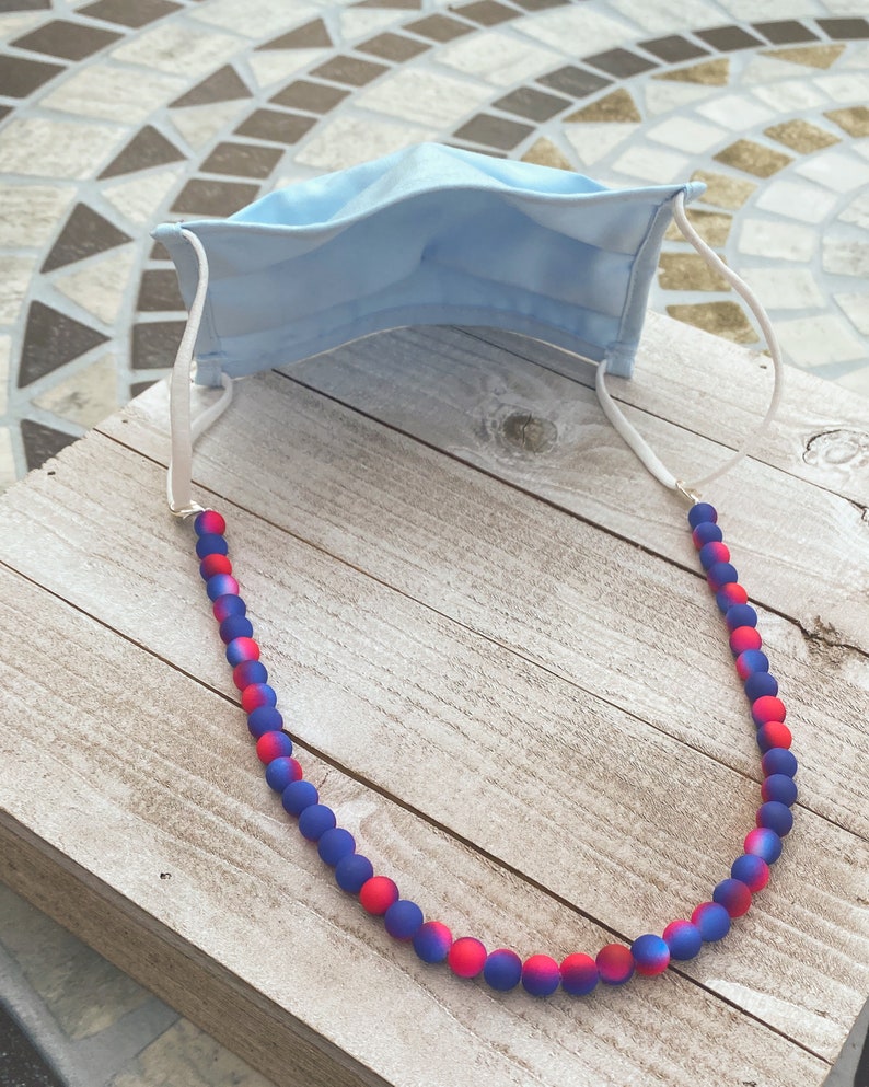 May include: A blue face mask with a white beaded chain. The beads are ombre red and blue.