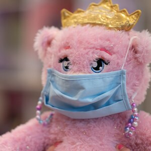 May include: A pink plush toy bear wearing a blue face mask and a gold crown. The bear has sparkly pink fur and large blue eyes. The mask has a light blue color and is secured with a beaded strap.