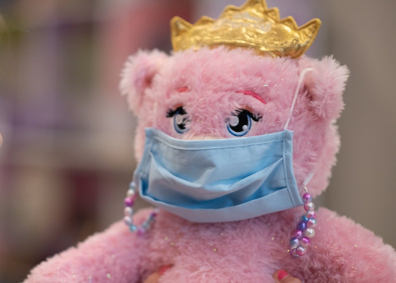 May include: A pink plush toy bear wearing a blue face mask and a gold crown. The bear has sparkly pink fur and large blue eyes. The mask has a light blue color and is secured with a beaded strap.