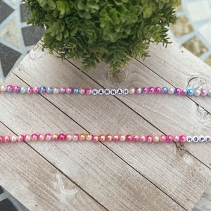 May include: Two beaded necklaces with the names "HANNAH" and "HOPE" spelled out in white letters. The beads are iridescent and come in shades of pink, blue, and orange.