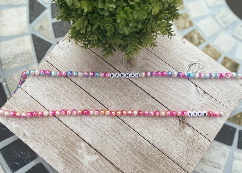 May include: Two beaded necklaces with the names "HANNAH" and "HOPE" spelled out in white letters. The beads are iridescent and come in shades of pink, blue, and orange.