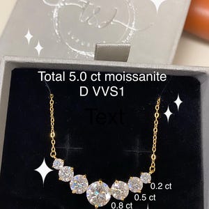May include: A gold chain necklace with a row of five round, clear moissanite stones. The stones are labeled with their carat weight: 2 ct, 0.8 ct, 0.5 ct, and 0.2 ct. The necklace is in a black velvet box with the text "Total 5.0 ct moissanite D VVS1" on the lid.