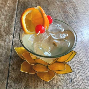 Può includere: Un cocktail trasparente in un bicchiere con ghiaccio, guarnito con una fetta d'arancia e due ciliegie rosse, servito in un bicchiere su un sottobicchiere a forma di loto dorato.