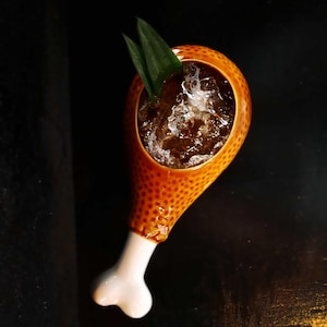Könnte beinhalten: Ein ausgefallenes Cocktailglas in Form einer Hähnchenkeule. Das orangefarbene Keramikglas hat eine strukturierte Oberfläche und einen weißen, knochenförmigen Sockel. Es enthält Eis, eine braune Flüssigkeit und ist mit zwei grünen Blättern garniert.