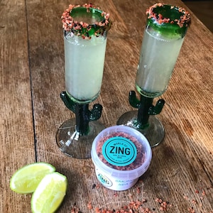 May include: Two green cactus-shaped shot glasses filled with a clear liquid and a red and brown rim. A small white container with a red and brown lid contains "World of Zing Smoked Sea Salt" with the text "Born in London 2014" and a lime wedge.