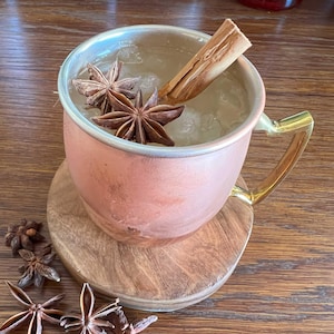 Puede incluir: Una taza de color cobre con un asa dorada, llena de un líquido transparente y hielo. Adornada con anís estrellado y una ramita de canela. La taza está sobre un posavasos de madera. La bebida está sobre una mesa de madera.