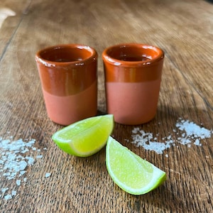 Puede incluir: Dos vasos de chupito de terracota con borde marrón y base naranja claro. Los vasos están sobre una superficie de madera con dos gajos de lima y sal blanca esparcida alrededor.