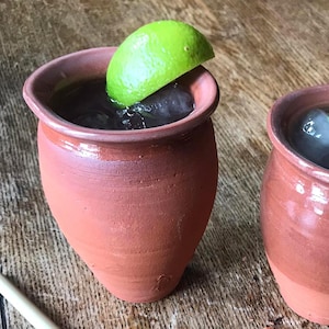 Puede incluir: Dos tazas de arcilla de color terracota, cada una con un líquido oscuro y hielo, adornadas con una rodaja de lima. Las tazas tienen un borde ancho y un cuerpo redondeado. Están sobre una superficie de madera.
