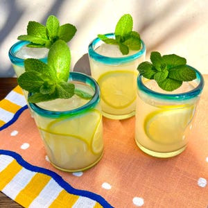Op de afbeelding: Vier glazen limonade met ijs, citroenschijfjes en muntblaadjes. De glazen hebben een turquoise rand en staan op een perzikkleurige doek met een geel-blauwe gestreepte rand. De scène wordt verlicht door zonlicht.