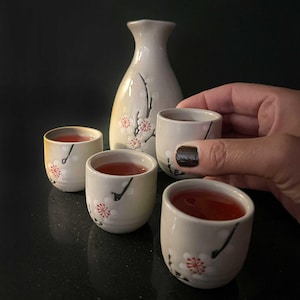 Puede incluir: Un juego de sake con una botella de cerámica y cuatro tazas pequeñas. El juego blanco roto está decorado con delicados diseños florales rojos y blancos y detalles en negro. Las tazas están llenas de un líquido rojo.