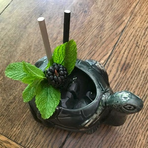 May include: A green ceramic turtle mug with a black blackberry and mint garnish. Two straws, one black and one white, stick out of the mug.