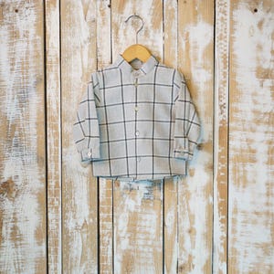 Può includere: Una camicia a maniche lunghe con bottoni in tessuto grigio chiaro con un motivo a griglia grigio scuro. La camicia presenta un colletto classico, un'abbottonatura frontale con bottoni chiari abbinati e polsini abbottonati, su una gruccia in legno.