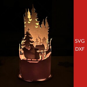 May include: A white paper lantern with a cut-out design of a winter scene featuring Santa Claus in his sleigh pulled by reindeer, a church, and trees. The lantern is lit from within, casting a warm glow.