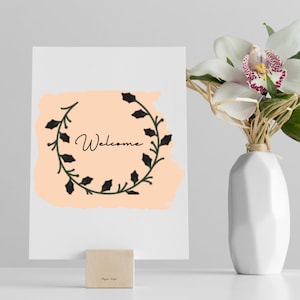 K&ouml;nnte beinhalten: Eine wei&szlig;e Karte mit einem pfirsichfarbenen Hintergrund und einem schwarzen floralen Kranz mit dem Wort "Willkommen" in schwarzer Schrift. Die Karte steht auf einem Holzblock auf einer wei&szlig;en Oberfl&auml;che. Eine wei&szlig;e Vase mit einer wei&szlig;en Orchidee befindet sich im Hintergrund.