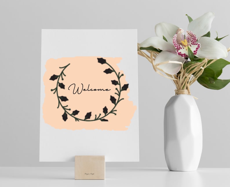 K&ouml;nnte beinhalten: Eine wei&szlig;e Karte mit einem pfirsichfarbenen Hintergrund und einem schwarzen floralen Kranz mit dem Wort "Willkommen" in schwarzer Schrift. Die Karte steht auf einem Holzblock auf einer wei&szlig;en Oberfl&auml;che. Eine wei&szlig;e Vase mit einer wei&szlig;en Orchidee befindet sich im Hintergrund.